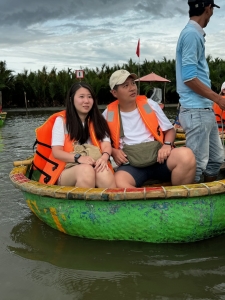Man and woman in orange life vests sit in round boat on water, guided by a person in blue shirt and hat.