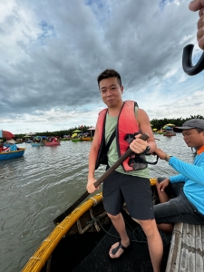 Man in red life vest holding paddle on small boat with others, calm water, cloudy sky, recreational.