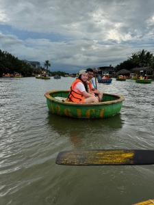 Two people in life jackets sit in a small green boat on a large water body with overcast skies.